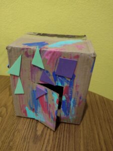 The front of a painted cardboard box house with foam shapes glued on it