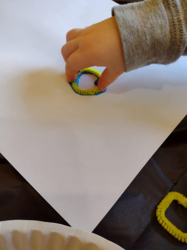 Picture of a preschooler placing a circle shaped pipe cleaner covered with paint onto a piece of paper