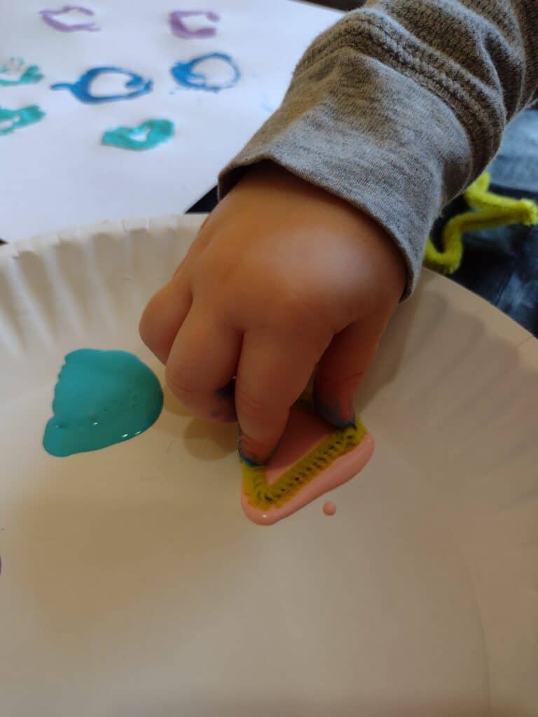 Picture of a preschooler putting a triangle shaped pipe cleaner in pink paint