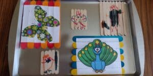 Picture of five popsicle stick puzzles on a cookie sheet. Three are drawn with markers and two are printed out pictures