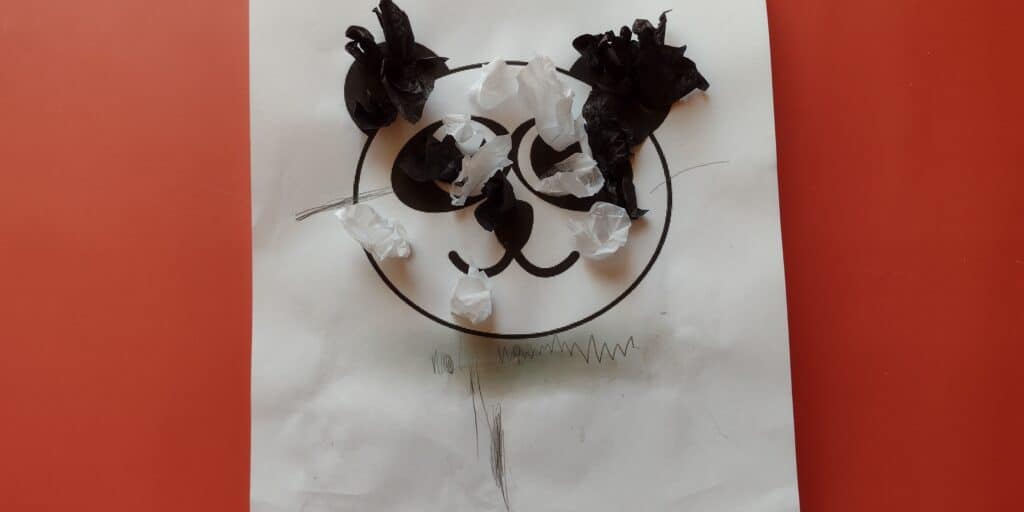 Picture of a 3d textured tissue paper panda craft made by a preschooler. There are only a few pieces of tissue paper glued to a picture of a panda and arms and legs are drawn on with pencil