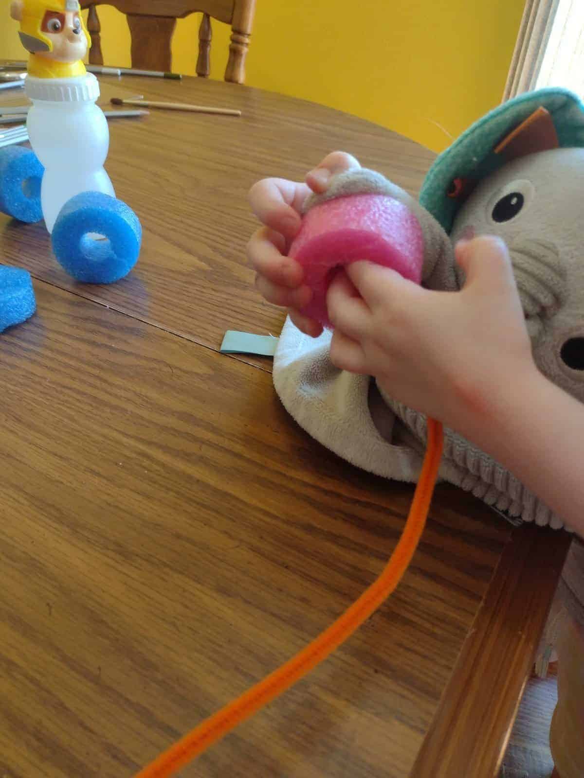 Picture of a preschooler using an elephant lovey to thread a piece of a pool noodle onto a pipe cleaner