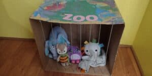 Picture of a pretend zoo made from a cardboard box. The bars are made from yarn. The box is painted and is labeled ZOO. Several stuffed animals are inside of it.