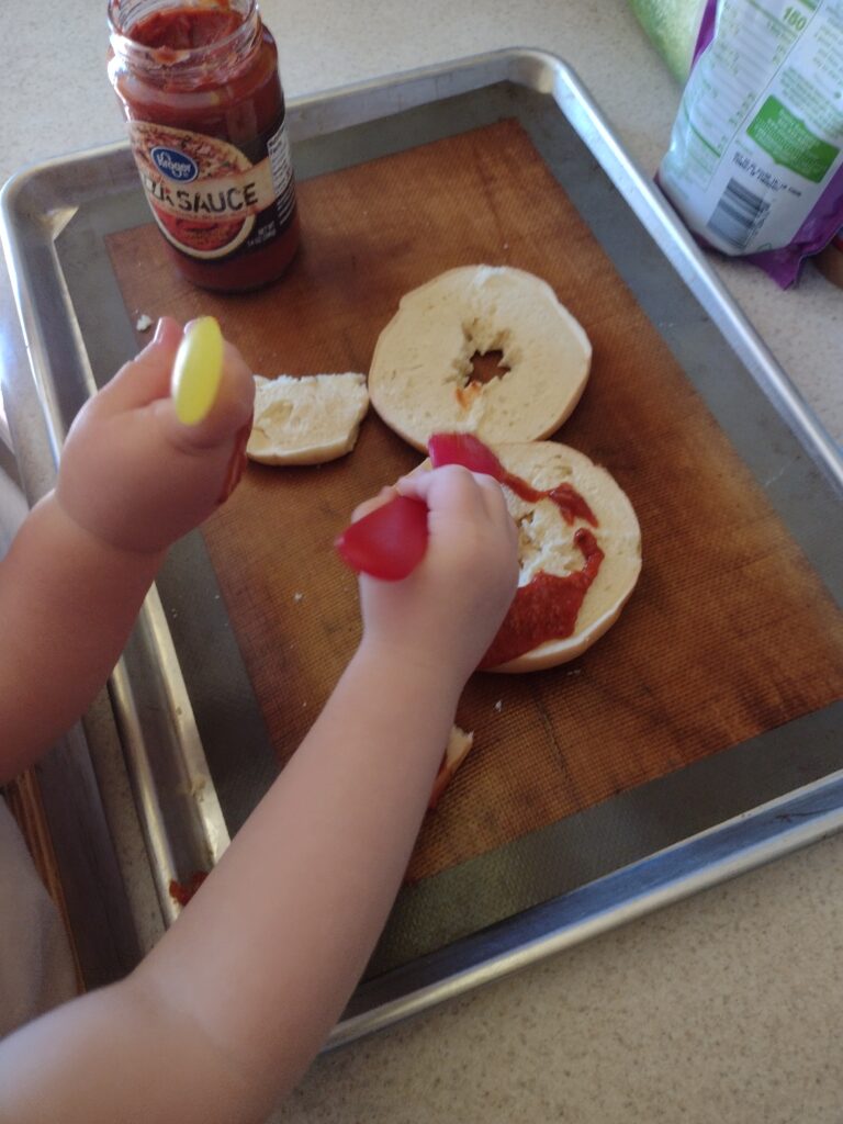 Picture of a preschooler and toddler spreading pizza sauce on bagels
