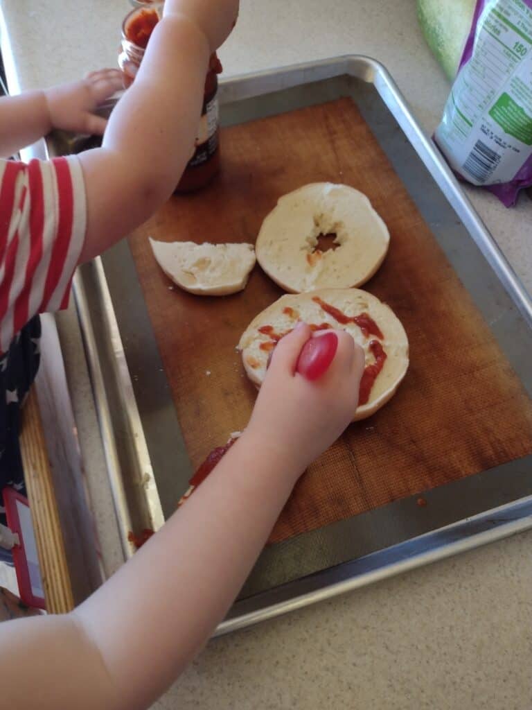Picture of a preschooler spreading pizza sauce on a bagel while a toddler gets pizza sauce on a spoon