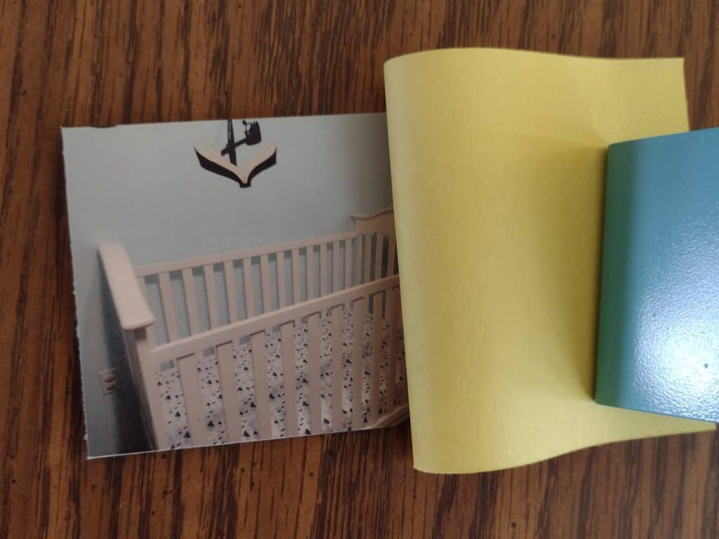 Picture of a picture of a crib taped facing a piece of yellow construction paper that is being held back to reveal the picture