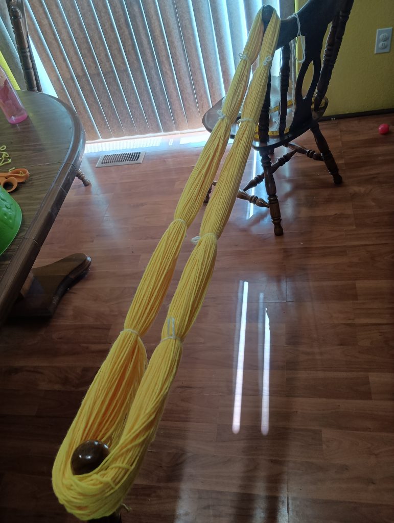 Picture of an entire skein of yellow yarn looped around two chairs and tied with white yarn every so often