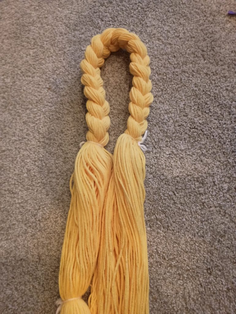 Picture of a large bunch of yellow yarn with a short braid in the middle of it. Loose yarn is hanging down from both sides of the braid