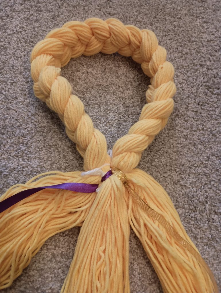 Picture of purple and gold ribbon tied to the top of yellow yarn about to be braided