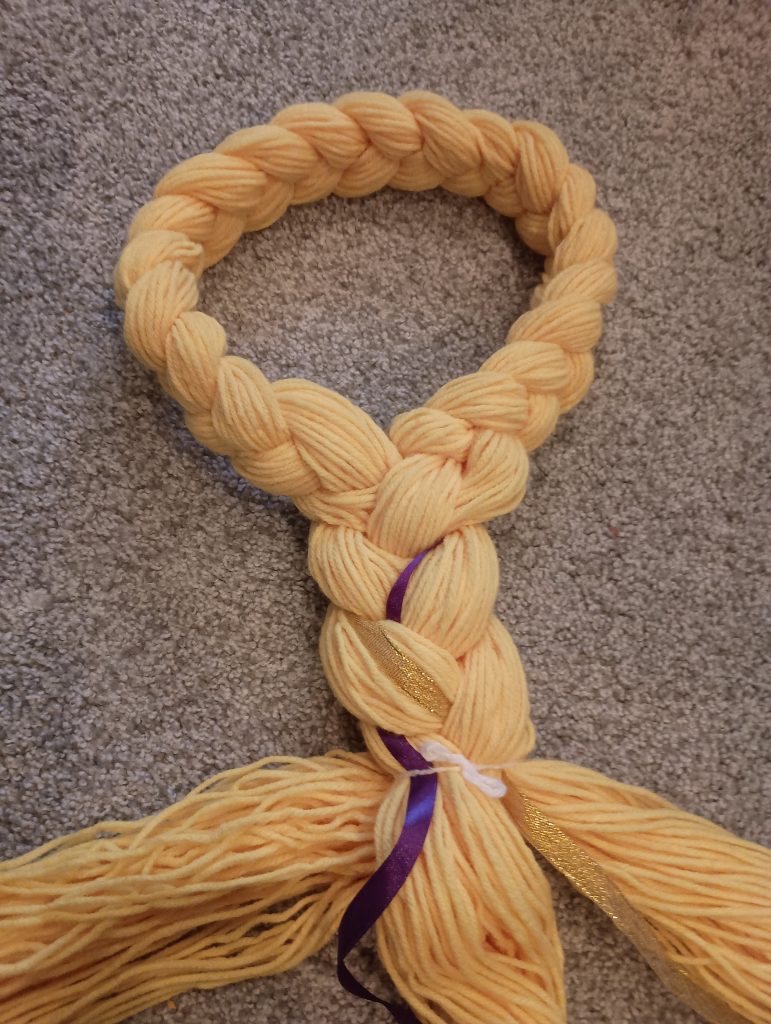 Picture of the start of braiding Rapunzel hair. A braided loop is on top, followed by a thicker braid of yellow yarn with ribbon braided with it and ending in three strands of loose yarn that still needs braided